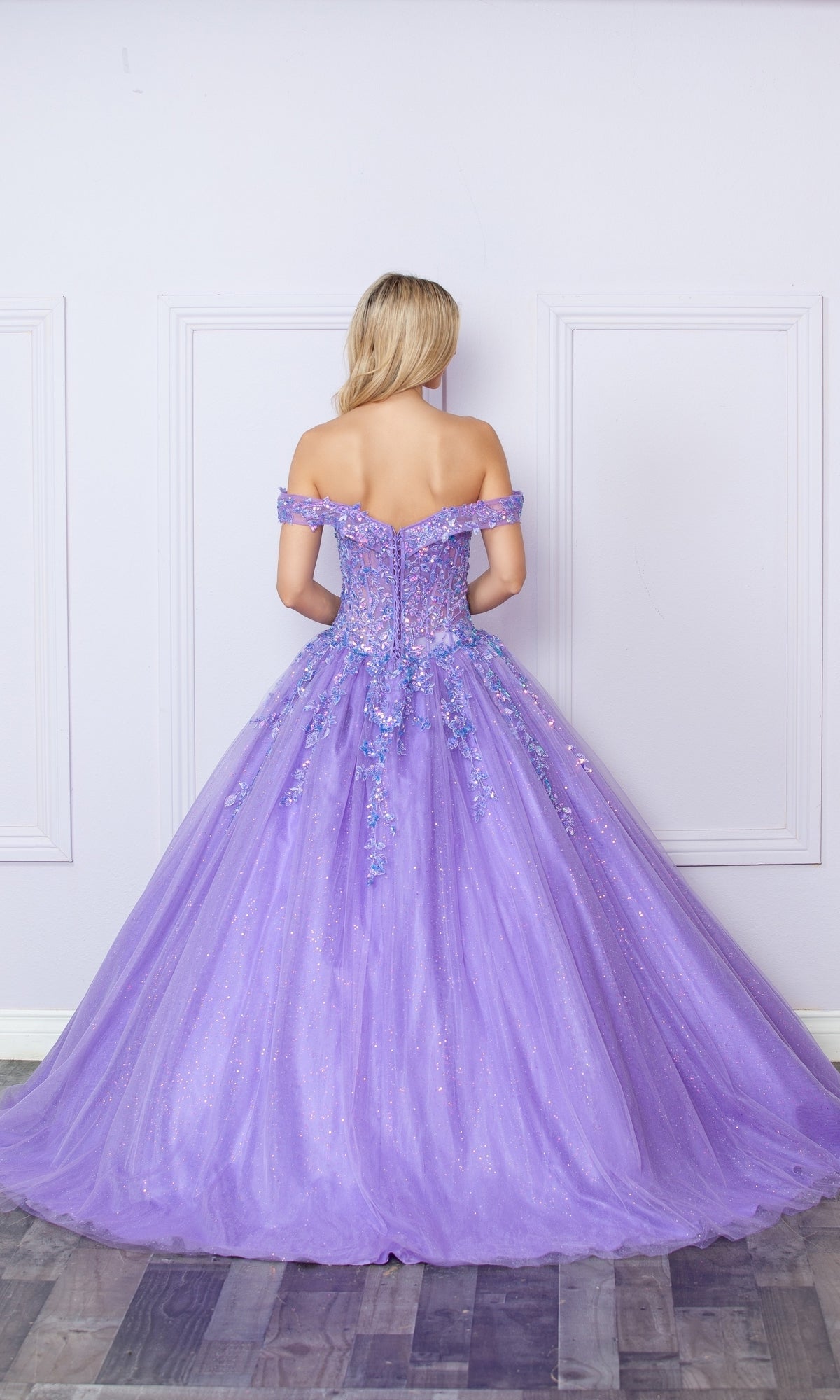 A woman faces away from the camera against a pale wall in the Bright Off-the-Shoulder Long Prom Ball Gown H1349, featuring a lavender hue, full tulle skirt, and floral embellishments.